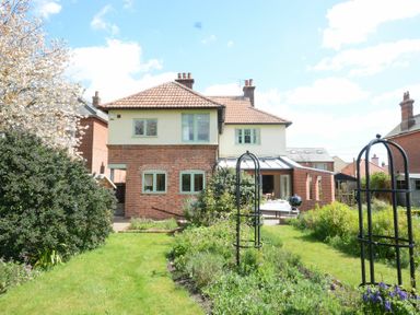 A backyard garden with grass and plants behind a two-story house at Holly Lodge in Aldeburgh