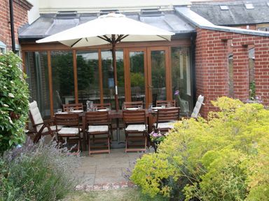 An outdoor patio with a wooden dining table and chairs under a large umbrella surrounded by garden plants at Holly Lodge in Aldeburgh