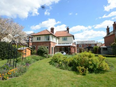 A backyard garden with a lawn flower beds and a two story house with a glass patio at Holly Lodge in Aldeburgh
