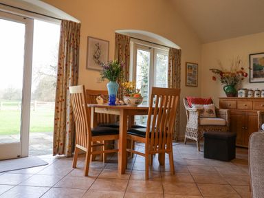 A dining area with a wooden table and chairs a wicker chair a wooden sideboard and flowers in a vase at Romulus in Larling near East Harling