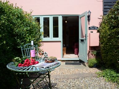 A garden patio area with a metal table and chair set a bottle of wine wine glasses roses and a newspaper outside a cottage with an open door at Seafarers in Fressingfield