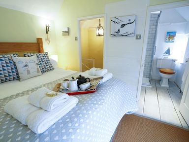 A bedroom with a bed with patterned pillows and folded towels on it with a tea set and bathroom visible at Aldeburgh Cottage in Aldeburgh