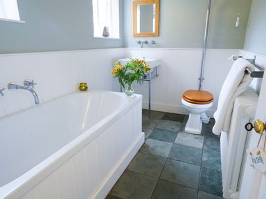 A bathroom with a bathtub flowers on the edge a small sink a toilet with a wooden lid and a towel hanging on a rail at Aldeburgh Cottage in Aldeburgh