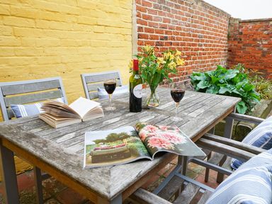 An outdoor wooden table with two glasses of red wine a wine bottle flowers an open book and an open magazine with striped cushions on wooden chairs at Aldeburgh Cottage in Aldeburgh