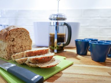 A kitchen counter with sliced bread on a cutting board a knife a French press with coffee and blue mugs