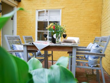 An outdoor patio with a wooden table and six chairs with pillows and a yellow brick wall at Aldeburgh Cottage in Aldeburgh