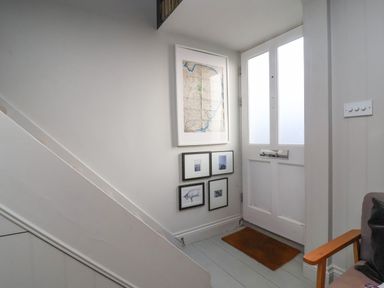 A hallway with framed pictures on the wall a wooden door with frosted glass panels a brown doormat and a wooden armchair at Mariners Cottage in Aldeburgh