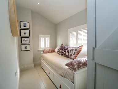 A small bedroom with a daybed with drawers under a window with shutters and framed pictures on the wall at Mariners Cottage in Aldeburgh