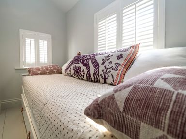A bed with patterned pillows and a polka dot sheet under windows with white shutters at Mariners Cottage in Aldeburgh