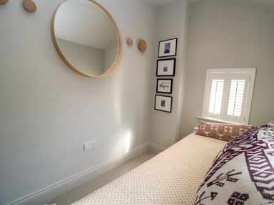 A bedroom with a bed near a window with shutters a round wall mirror and framed pictures on the wall at Mariners Cottage in Aldeburgh