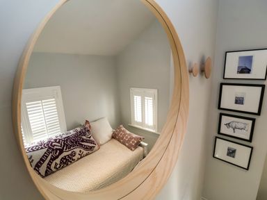 A bed with three pillows reflected in a round mirror with four framed pictures hanging on a wall at Mariners Cottage in Aldeburgh