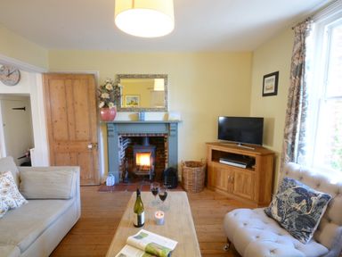 A living room with a fireplace a wooden door a sofa a TV on a wooden cabinet and a coffee table with wine glasses at Pebble Beach Cottage Aldeburgh