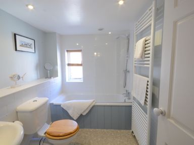 A bathroom with a bathtub a toilet with wooden seat a towel rack and a small window at Pebble Beach Cottage Aldeburgh