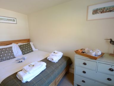 A bedroom with a double bed with cushions and folded towels and a dresser with a tea set and a decorative bird at Pebble Beach Cottage Aldeburgh