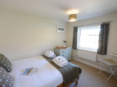 A bedroom with a bed with folded towels on it a dresser with a tray and a lamp a window with curtains and a chair by the window at Pebble Beach Cottage Aldeburgh