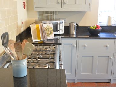 A kitchen countertop with a gas stove a utensil holder a microwave kettle an open cookbook and a bowl of apples at Pebble Beach Cottage Aldeburgh