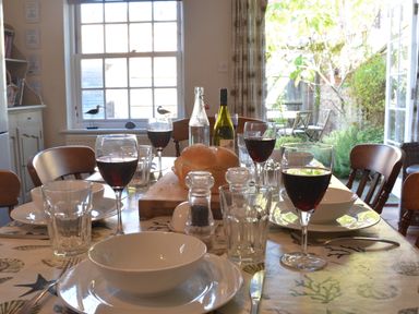 A dining table set with bowls plates glasses and wine glasses with bread on a cutting board in a kitchen with an open door to a garden at Pebble Beach Cottage Aldeburgh