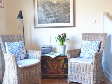 Two wicker chairs with patterned cushions around a decorated wooden chest with a plant and radio on top at Pebble Beach Cottage Aldeburgh