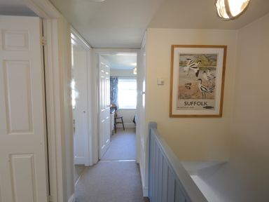 A hallway with doors leading to rooms a framed Suffolk poster on the wall and a blue staircase railing at Pebble Beach Cottage Aldeburgh