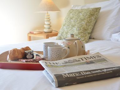 A breakfast tray with croissants jam cups and a teapot on a bed with a pillow and a newspaper at Pebble Beach Cottage Aldeburgh