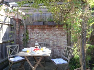 An outdoor patio with a wooden table and three chairs under a pergola with climbing plants at Pebble Beach Cottage Aldeburgh