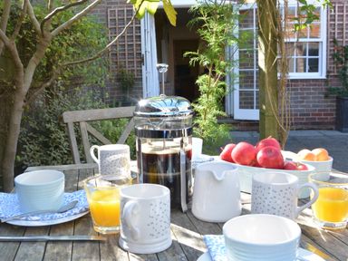 A wooden outdoor table with coffee, orange juice, cups, and bowls of fruit at Pebble Beach Cottage Aldeburgh