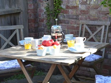 An outdoor wooden table with cups glasses bowls of fruit and a coffee press with wooden chairs at Pebble Beach Cottage Aldeburgh