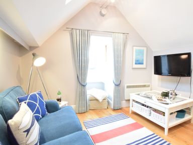 A small living room with a blue sofa pillows and floor lamp near a window with curtains a wall mounted tv and a white coffee table with magazines and baskets at Pied a Mer Aldeburgh