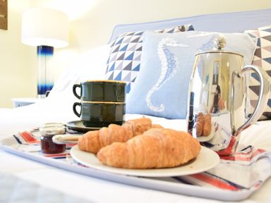 A tray with croissants, two stacked cups, a small jar and a metallic coffee pot on a bed with pillows including one with a seahorse design at Pied a Mer, Aldeburgh in Aldeburgh