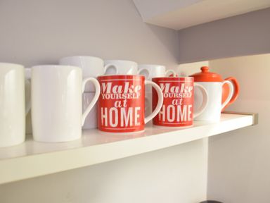 A kitchen shelf with white mugs and red mugs that say Make Yourself at Home and a white and red teapot at Pied a Mer Aldeburgh in Aldeburgh