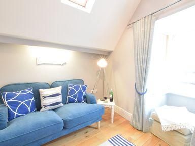 A blue sofa with cushions next to a small white table with a bottle and glass of wine and a floor lamp in a living room at Pied a Mer Aldeburgh