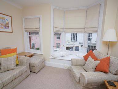 A sitting room with two plaid sofas orange and yellow cushions a bay window with cushions and beige blinds a floor lamp and wooden side tables at The Hideaway Aldeburgh