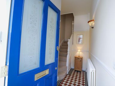 An entrance hallway with a blue front door an upright small wooden cabinet with a lamp a staircase with carpet a radiator and a framed picture on the wall at The Hideaway in Aldeburgh