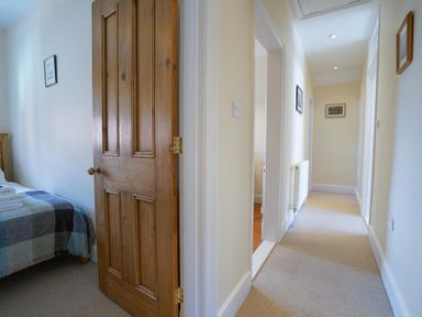 A hallway with carpeted floor and framed pictures on walls with an open wooden door leading to a bedroom at The Hideaway in Aldeburgh