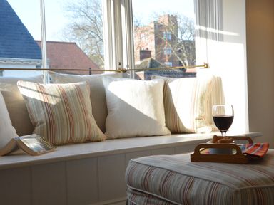 A window seat with striped and plain cushions and a book next to a footstool with a tray holding a glass of red wine at The Hideaway in Aldeburgh