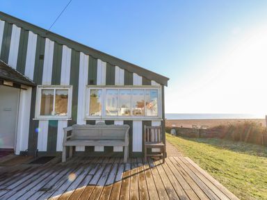 A wooden deck with a bench and chair outside a striped cabin near a grassy area and a beach at Valetta Thorpeness in Thorpeness