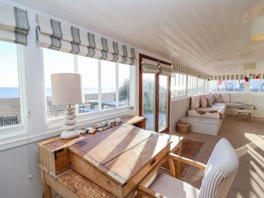 A room with a wooden desk, a lamp, a striped chair, a sectional sofa with cushions, and windows showing a beach view at Valetta, Thorpeness in Thorpeness