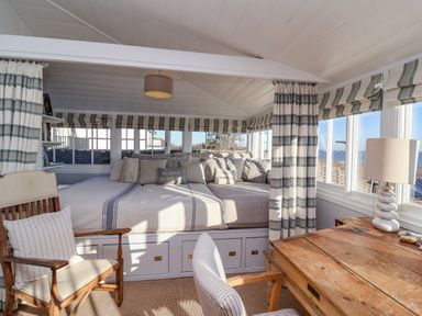 A room with a wooden desk and chair a built in sofa with cushions and large windows with striped curtains at Valetta Thorpeness in Thorpeness