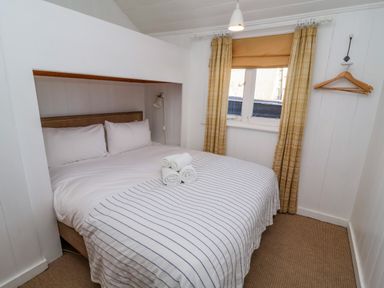 A bedroom with a double bed, white and striped bedding, three rolled towels on the bed, window with curtains, and wooden hangers on the wall at Valetta, Thorpeness