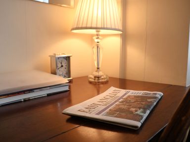 A wooden desk with a folded newspaper a closed folder a small clock and a lit table lamp at Winkle Cottage Aldeburgh