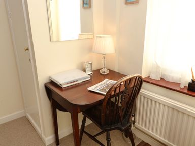 A small wooden desk with a chair a lamp a clock and a newspaper by a window at Winkle Cottage Aldeburgh