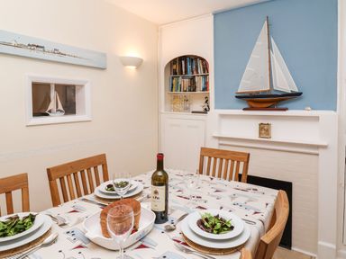 A dining room with a table set for four including bowls of salad wine bottle and bread with wooden chairs model sailboats and bookshelf in the background at Winkle Cottage Aldeburgh