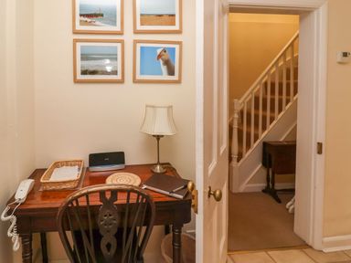 A small desk with a chair lamp phone and framed pictures above near a doorway showing stairs at Winkle Cottage Aldeburgh