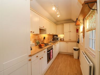 A kitchen with white cabinets wooden countertops a stove an oven a microwave a toaster a fruit bowl and windows with floral curtains at Winkle Cottage Aldeburgh