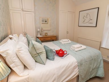 A bedroom with a bed featuring multiple pillows and folded towels on top a side table with a fan and a clock wardrobes and a wall picture of a bird at Winkle Cottage Aldeburgh