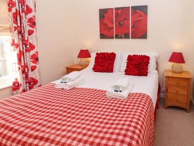A bedroom with a bed covered by a red and white checkered blanket and red floral pillows and curtains at Winkle Cottage Aldeburgh