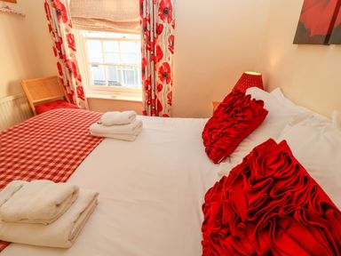 A bedroom with a bed with red and white bedding near a window with red floral curtains and a wooden chair at Winkle Cottage Aldeburgh