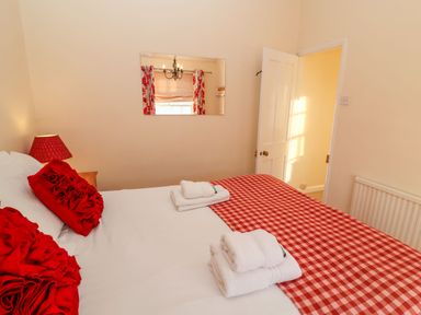 A bedroom with a bed with red and white checkered bedding and red pillows next to a wooden nightstand with a red lamp and a mirror on the wall at Winkle Cottage Aldeburgh