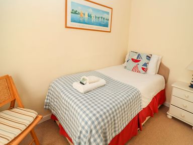 A bedroom with a single bed covered in a blue check blanket and red skirt with a pillow and folded towels on the bed a wooden chair with striped cushion a white nightstand with drawers and a lamp a framed picture on the wall at Winkle Cottage Aldeburgh
