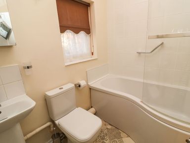 A bathroom with a toilet, sink, bathtub with shower screen, and a window with a brown blind at Winkle Cottage Aldeburgh